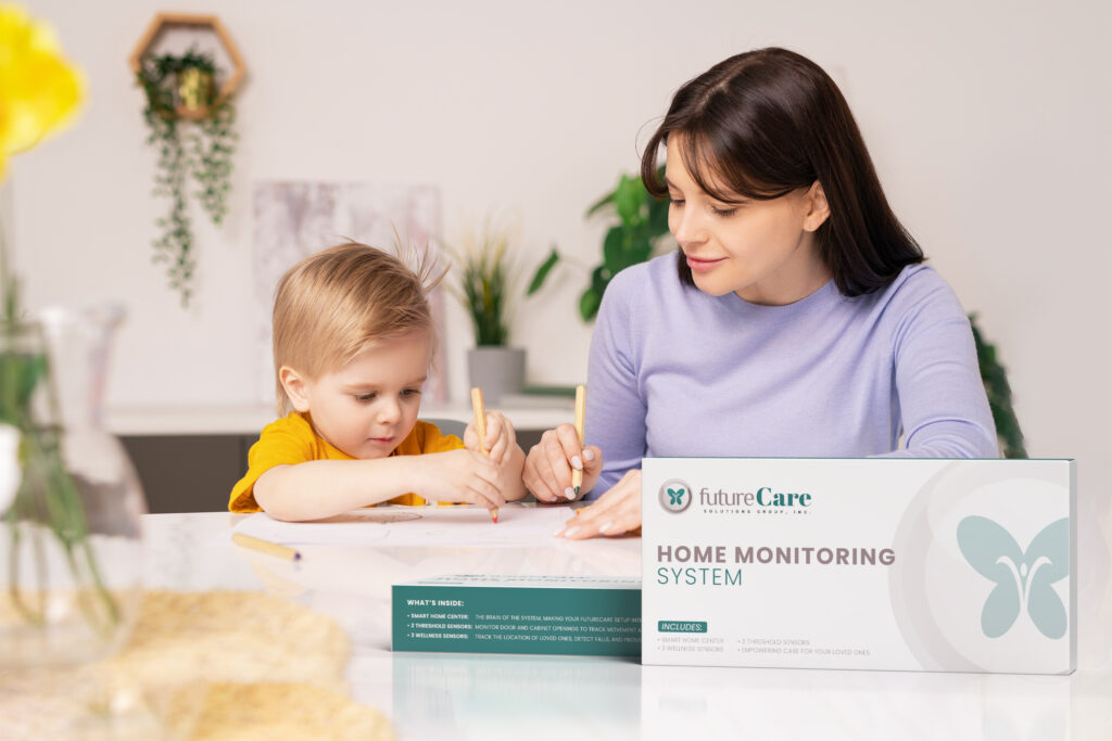 Mother and young child sit at a white table drawing together, with a FutureCare Home Monitoring System box in the foreground.